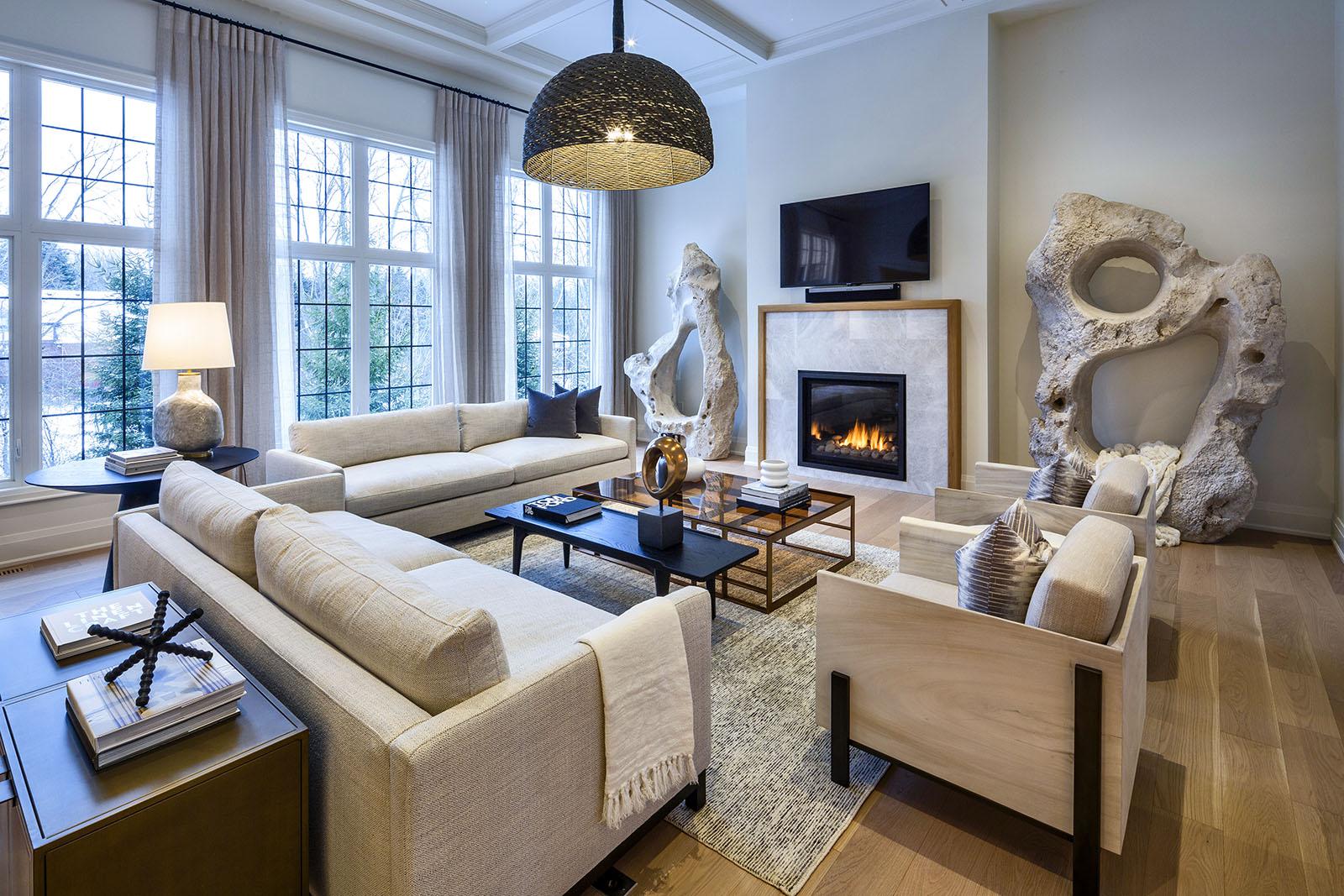 Image of a beige-themed living room with comfy sofas, a fireplace, rocky decorative pieces, and large windows letting in soft natural light.